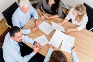 Overhead view of several professionals around a wooden table reviewing and signing documents.]","altTexts