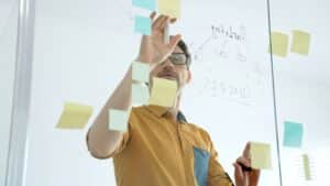 Person wearing glasses places colorful sticky notes on a glass wall during a brainstorming session.