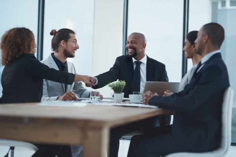 Business colleagues in a meeting shake hands across a conference table, smiling after an agreement.