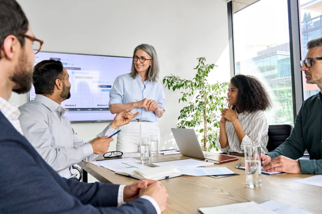 Group of businesspeople in a modern office, a woman presenting beside a screen with charts on it.