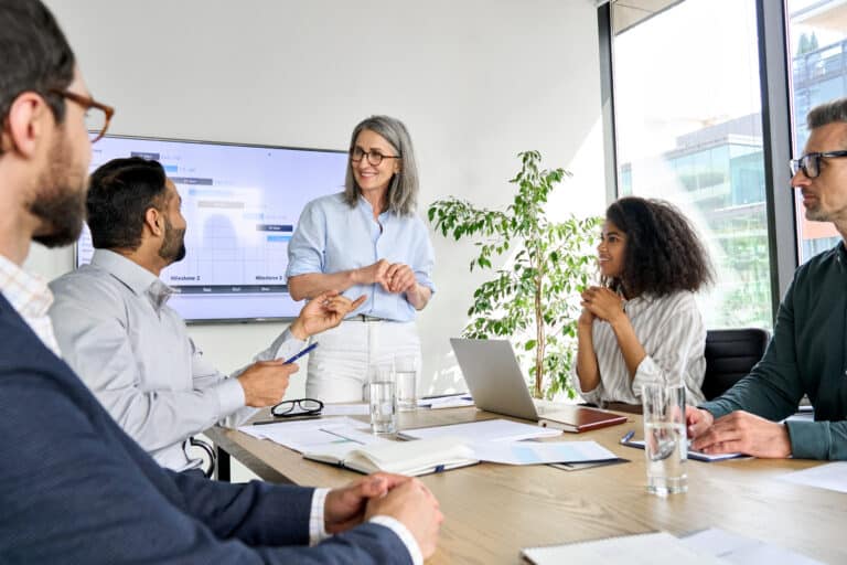 Group of businesspeople in a modern office, a woman presenting beside a screen with charts on it.