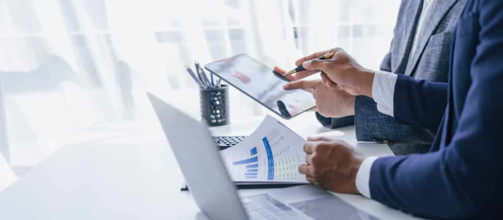 Two professionals in business attire review a tablet and printed charts at a bright office desk, analyzing data.